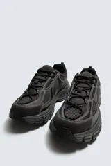 Zapatilla deportiva de estilo running, completamente negra, con capellada de malla y superposiciones sintéticas. Presenta una suela gruesa y voluminosa con diseño segmentado.
