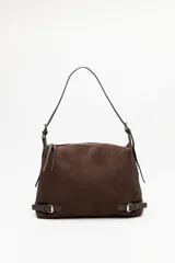 Cartera de gamuza color marrón, de textura suave y acabado aterciopelado. Cuenta con una silueta amplia y flexible, cierre superior metálico, correa regulable y detalles laterales con hebillas.