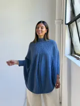 Poncho de tejido de punto color azul jean, con cuello alto y diseño de trenzas verticales. Presenta mangas integradas con puños acanalados y corte holgado.