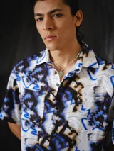 Camisa de manga corta de calce relajado, con cuello clásico, cierre frontal con botones y bolsillos aplicados en el pecho. Presenta un estampado integral abstracto en tonos crudo, negro, marrón y azul eléctrico.
