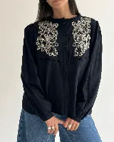 Camisa de color marrón chocolate con bordado artesanal de flores y hojas en hilo crudo. Presenta cuello con volado, cierre frontal de botones y mangas largas con puños abotonados.