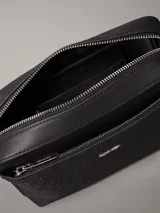 Cartera tipo cámara convertible Calvin Klein negra, con textura monograma en el bolsillo frontal y logo metálico plateado. Incluye dos correas desmontables: una ancha ajustable para cruzar y una fina para llevar al hombro.