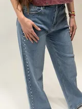 Jean de denim azul de corte recto, con detalles de tachas metálicas plateadas en los bolsillos delanteros y a lo largo de la costura exterior de la pierna derecha.