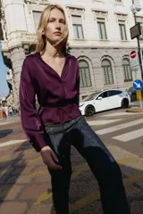 Camisa satinada de color violeta oscuro, con cuello solapa, escote en pico y manga larga. Presenta un detalle de cinturón fino ajustable a tono en la cintura y cierre frontal con botones.