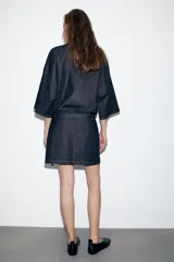 Vestido corto de denim azul oscuro, con cuello redondo, mangas amplias de largo tres cuartos y cintura elástica con cinturón integrado del mismo tejido. Presenta costuras a contraste y cierre posterior con cremallera oculta.