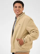 Campera bomber reversible azul marino y beige con cuello, puños y ruedo acanalados con rayas doradas. Tiene cierre frontal y logo bordado en el pecho.