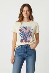 Remera de algodón color crema con cuello redondo y manga corta. Presenta un estampado artístico de un ramo de flores en tonos rosados, azules y morados, dispuesto en un jarrón sobre una superficie.