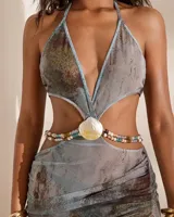 Vestido corto con diseño cut-out en la cintura, escote halter y detalle central de concha marina con cuentas de colores. Presenta un estampado abstracto en tonos azulados y verdosos.