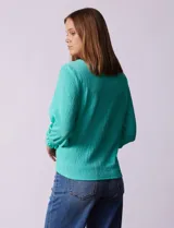 Blusa blanca de tejido texturado con cuello redondo, escote en V con aplique metálico y mangas 3/4 con puños elásticos. El ruedo se ata con un nudo.