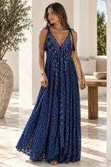 Vestido largo color dorado, confeccionado en tela semitransparente con diseño bordado floral y detalles de brillo. Presenta escote en V, tirantes finos ajustables con borlas en los hombros, y corte amplio y fluido con gran movimiento.