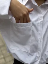 Camisa blanca de manga larga con sutiles rayas verticales, cuello camisero, cierre frontal con botones y dos bolsillos frontales con solapa.