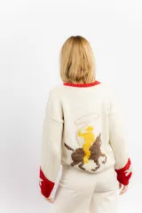 Sweater tejido a mano en lana merino color crudo, con cuello y puños en rojo. Presenta un diseño con motivos de un cowboy y estrellas.