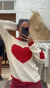 Sweater de punto de lana merino en color crudo, con diseño de corazón grande en color rojo en el centro y detalles de corazones en las mangas. Presenta cuello redondo, puños y ruedo acanalados en color rojo.