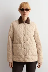 Campera capitoneada de diseño acolchado con patrón de rombos, color beige claro. Presenta cuello camisero de pana en color marrón oscuro, cierre frontal con botones metálicos y bolsillos frontales de parche.