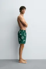 Bañador de hombre largo, color verde oscuro, con estampado de hojas blancas. Presenta cintura ajustable con cordón beige y forro interior.