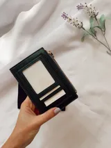 Tarjetero de diseño rectangular con cierre superior, confeccionado en material texturizado color verde con detalles en cuero sintético. Incluye el nombre de la marca grabado en el frente.