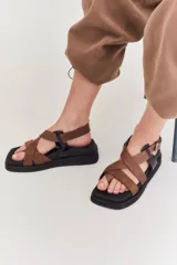 Sandalias de plataforma plana con diseño de tiras cruzadas en tonos beige y marrón. Presentan una base negra, puntera cuadrada y cierre con hebilla metálica lateral.