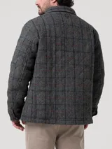 Chaqueta de hombre con diseño acolchado, confeccionada en lana Harris Tweed con patrón de tartán en tonos grises. Presenta cuello camisero, cierre frontal con botones de imitación cuero, bolsillos frontales y relleno térmico de fibras recicladas.