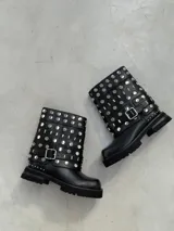 Bota corta de cuero negro con suela track, plataforma baja y taco cuadrado. Está decorada con múltiples tachas metálicas plateadas en toda la caña y cuenta con una tira con hebilla lateral en el tobillo.