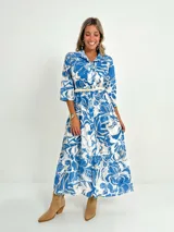 Vestido camisero midi de algodón con estampado floral en tonos rosados y blancos. Tiene cuello camisero, mangas 3/4, botones frontales, falda escalonada y cinturón fino.