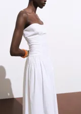 Vestido midi color blanco con diseño evasé, escote palabra de honor y sin mangas. Cuenta con detalles fruncidos en los laterales y cierre de cremallera en la parte posterior.