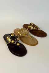 Sandalia plana de gamuza marrón, estilo ojota, con apliques metálicos dorados y hebilla decorativa.