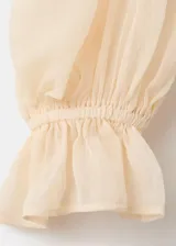 Camisa de diseño recto en tono beige, confeccionada con tejido combinado y panel de encaje frontal. Presenta cuello redondo, mangas largas abullonadas con puños elásticos y detalle de volantes en la cintura.