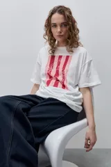 Remera blanca de calce relajado y silueta amplia, con cuello redondo, mangas cortas y estampa gráfica frontal de un cangrejo sobre franjas en tonos rosado y rojo.