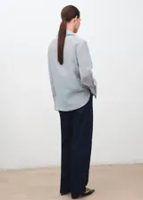 Blusa de tejido ligero color celeste con cuello camisero y cierre frontal de botones ocultos. Presenta mangas largas con puños anchos y detalles florales bordados en la parte superior delantera.