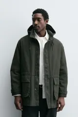 Parka color verde militar de corte relaxed fit, confeccionada en tejido de algodón con interior acolchado. Cuenta con cuello subido, capucha ajustable con cordón, cierre frontal de cremallera oculta por solapa con botones a presión y amplios bolsillos frontales de plastrón con solapa.
