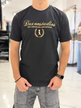 Remera color beige con cuello redondo y mangas cortas. Estampado frontal con el texto "Duxonstudios" en tipografía cursiva, acompañado de un logotipo circular con hojas de laurel y una figura central en tono verde oscuro.