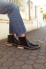 Botas tipo Chelsea de cuero negro, con paneles laterales elásticos, detalle de textura en el talón y suela plana con detalle en madera clara.