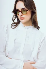 Camisa blanca de manga larga con cuello alto con volados, detalles de alforzas y pliegues en el frente y cierre frontal con botones ocultos.