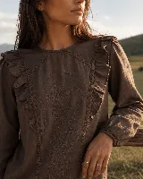 Blusa de color beige con bordados florales en el frente, escote redondo y mangas largas con puños elastizados. Presenta volados decorativos a los lados del pecho.