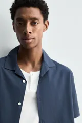 Camisa de manga corta con cuello bowling, confeccionada en popelín de algodón. Presenta un corte relajado, cierre frontal con botones y aberturas laterales en el bajo.