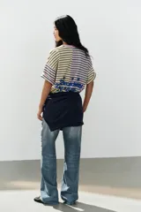 Camiseta oversize con diseño bicolor a rayas horizontales, combinando un lado en tonos rosado y verde oscuro, y el otro en beige y marrón. Cuenta con cuello redondo, mangas cortas amplias y texto bordado en azul en el pecho.