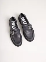 Mocasines de cuero gris oscuro con suela chunky, costuras visibles en la puntera y una tira superior ajustable con hebilla metálica plateada.