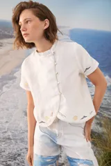 Blusa blanca de lino con mangas cortas, cuello alto con volado y cierre lateral cruzado con botones dorados. Presenta detalles bordados artesanales a contratono en el ruedo y las mangas.