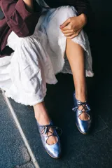 Zapato tipo blucher de piel metalizada en color violeta, con diseño híbrido entre blucher y bailarina. Presenta cierre con cordones y borlas decorativas, acabado sin ribete con ondas laterales y elástico en la parte trasera para un ajuste óptimo. Interior y suela de cuero.