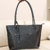 Cartera tipo tote de cuero marrón con costuras artesanales visibles, bolsillo frontal con el logo grabado, asas de hombro y detalles de flecos colgantes.