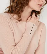 Suéter de punto color rosado, con cuello barco, mangas largas y puños con botones decorativos. El dobladillo también es acanalado.