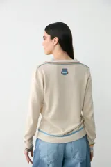 Remera de punto color crudo con cuello redondo y detalles de rayas en azul, amarillo y bordó. Presenta un parche bordado en el pecho con el logo de Manos del Uruguay y la inscripción 2026.