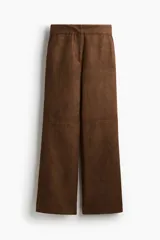 Pantalón de corte bootcut confeccionado en gamuza color marrón. Presenta cierre frontal con botón oculto, bolsillos laterales al bies y costuras decorativas horizontales sobre las rodillas.