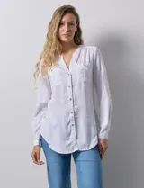 Camisa blanca de lino y rayón, con cuello a la base y escote en V abotonado. Tiene dos bolsillos con botón en el pecho y mangas largas regulables con presilla y botón.