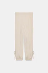 Pantalón de mujer color beige claro, de tiro medio y calce holgado, confeccionado en tejido ligero y fluido. Presenta cintura elástica ajustable con lazada, bolsillos laterales y aberturas ajustables con lazadas en las piernas que terminan en bajo abullonado con elástico.