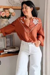 Camisa de color terracota con cuello camisero, cierre frontal de botones y mangas largas abullonadas. Presenta un delicado bordado floral en color blanco en la zona del pecho y los hombros, con un diseño de corte holgado.