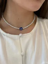 Collar corto de cuentas de cristal facetado en color blanco, con un dije central de ojo turco en tonos azul, blanco y negro.