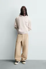Sudadera hoodie relaxed fit de color beige, con cuello con capucha, manga larga y bolsillo frontal tipo canguro. Presenta un pequeño estampado de texto en contraste en el pecho.
