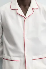 Pijama de dos piezas confeccionado en popelín de algodón blanco, con vivos de ribete en color bordó. Incluye una camisa de manga larga con cuello bowling, bolsillos de plastrón y cierre frontal de botones, y un pantalón de corte relajado con cintura elástica y bolsillos laterales.