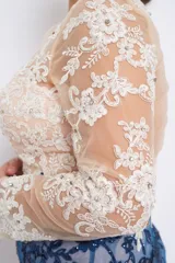 Vestido corto de fiesta con falda azul y parte superior color piel, ambos con bordado floral. El vestido tiene mangas largas transparentes con bordados y está confeccionado en tul con hilo y canutillos.